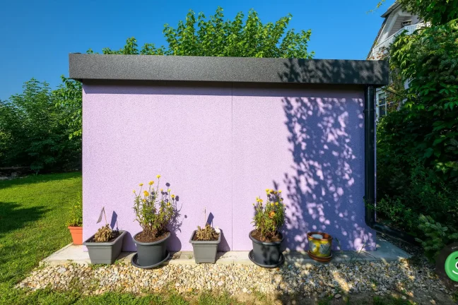 Záhradný domček s fialovou omietkou obklopený rôznymi kvietkami