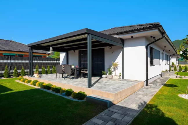 Antracitová pergola pri bielom dome s dláždenou terasou a záhradným sedením, modrá obloha
