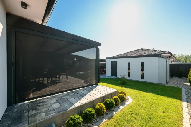 Antracitová pergola so zatiahnutou roletou pri bielom dome, dláždená terasa a zelený trávnik