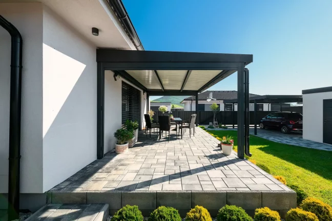 Antracitová pergola s lamelovou strechou pri bielom dome, dláždená terasa so záhradným sedením