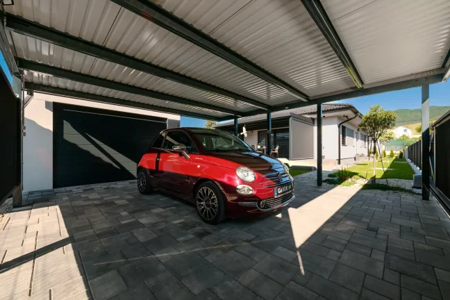 Červené auto pod prístreškom s pultovou strechou, pohľad z boku, dláždená plocha a zelená záhrada