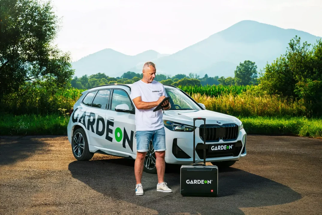 Tablet v rukách agenta GARDEON, firemné BMW a kufrík v prírode.