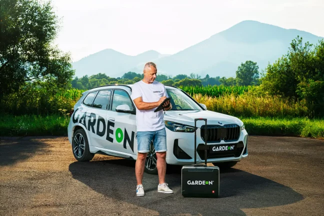 Tablet v rukách agenta GARDEON, firemné BMW a kufrík v prírode.