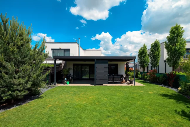 Antracitová pergola s plochou strechou pri bielom dome, rozľahlý zelený trávnik a stromy