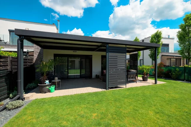 Voľne stojaca antracitová pergola s plochou strechou na zelenej záhrade, modrá obloha s oblakmi