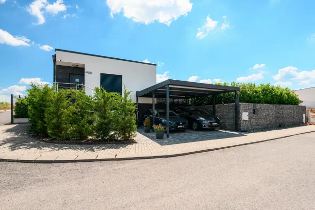 Antracitový prístrešok s plochou strechou pri rodinnom dome, ktorý kryje dve autá.