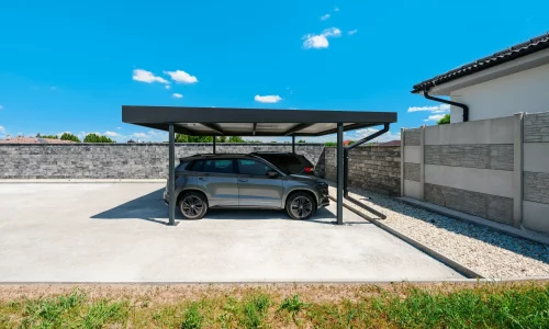 Dve SUV zaparkované pod antracitovým prístreškom s plochou strechou pri dome.