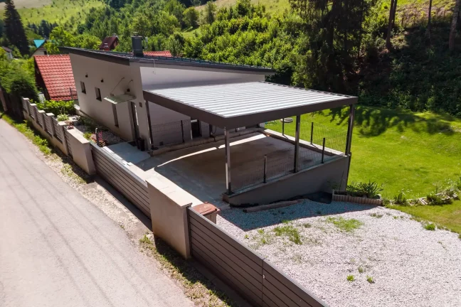 Vzdušný pohľad na prístrešok s trapézovou strechou pri bielom rodinnom dome.