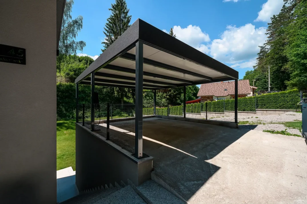 Tmavosivý prístrešok s plochou strechou ukončenou atikou pri rodinnom dome.