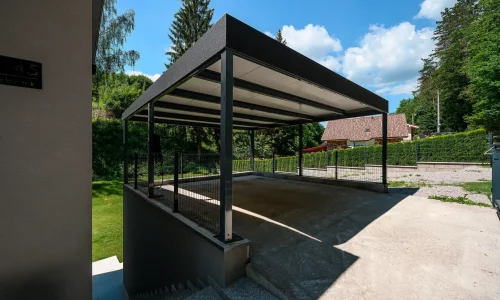 Tmavosivý prístrešok s plochou strechou ukončenou atikou pri rodinnom dome.