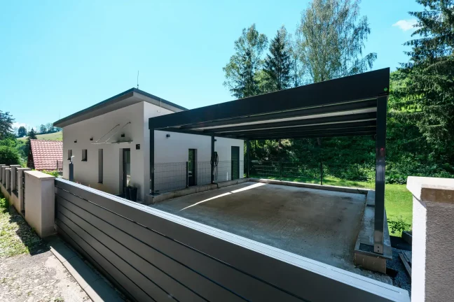 Otvorený prístrešok s antracitovou plochou strechou pri bielom rodinnom dome.