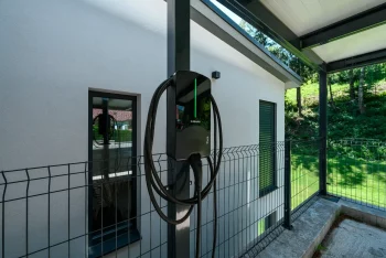 Nabíjačka elektromobilov na stĺpiku antracitového prístrešku pri omietanej fasáde.