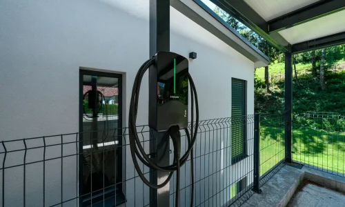 Nabíjačka elektromobilov na stĺpiku antracitového prístrešku pri omietanej fasáde.