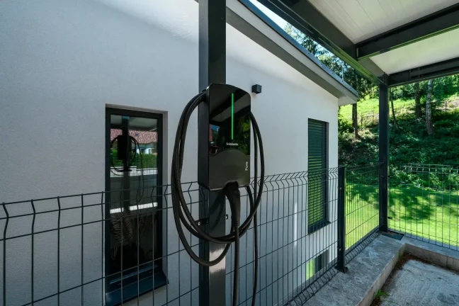 Nabíjačka elektromobilov na stĺpiku antracitového prístrešku pri omietanej fasáde.