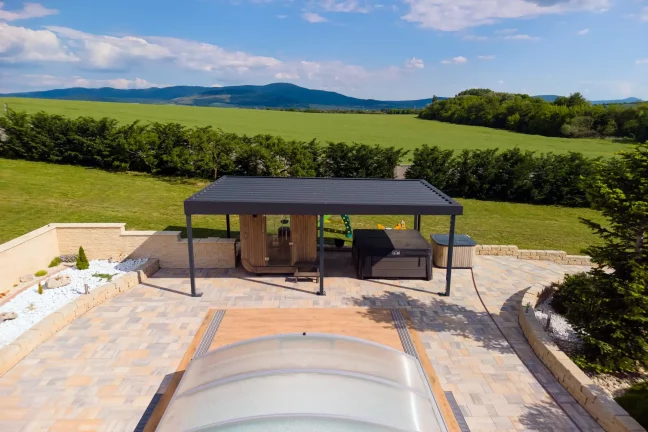 Antracitová pergola so záhradným sedením pri bazéne s priehľadným krytom, zelené kopce v pozadí