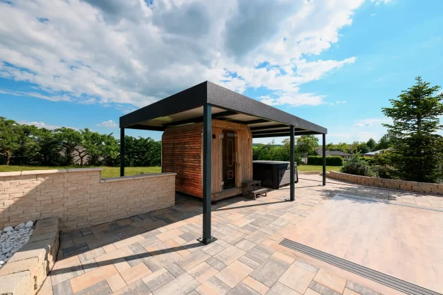 Voľne stojaca antracitová pergola s plochou strechou na dláždenej terase, zelená záhrada v pozadí
