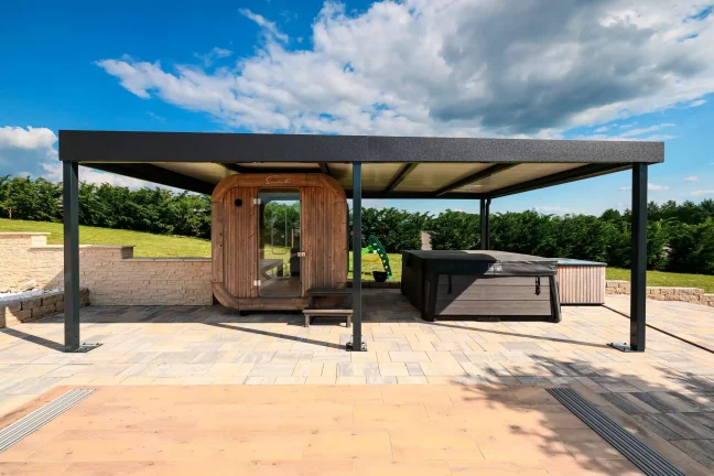 Antracitová pergola s plochou strechou pri sudovej saune na dláždenej terase, modrá obloha