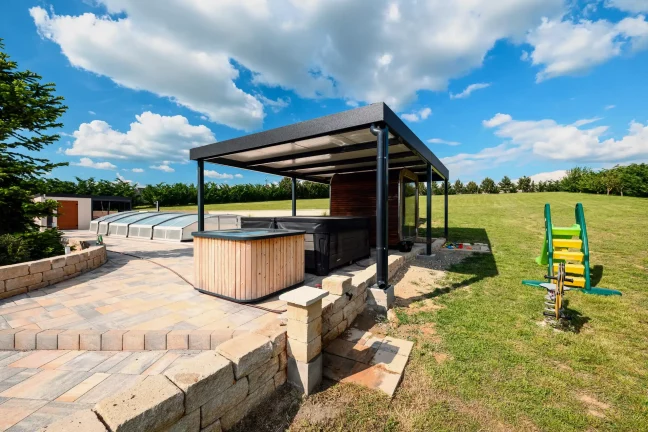 Antracitová pergola s vírivkou a detským ihriskom v pozadí, dláždená terasa pri záhrade