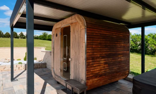 Sudová sauna z tmavého dreva pod antracitovou pergolou, dláždená plocha a zelený trávnik