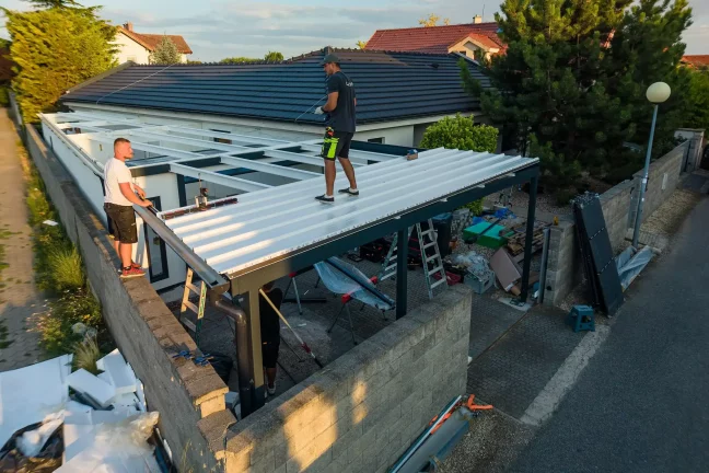 Dvaja montážnici chodia po bielej streche prístrešku nad kamenným múrikom.