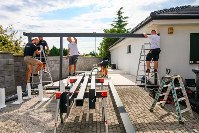 Montážnici zdvíhajú oceľový nosník rebríkmi pri stene domu