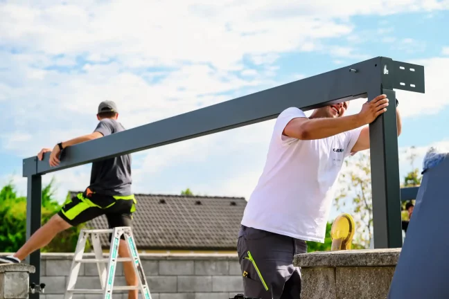 Montážnici spájajú oceľové nosníky rohovej konštrukcie prístrešku, jeden na rebríku.
