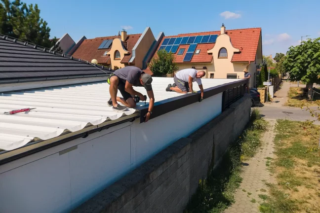Dvaja pracovníci kľačia na streche prístrešku a vŕtajú okrajovú lištu.