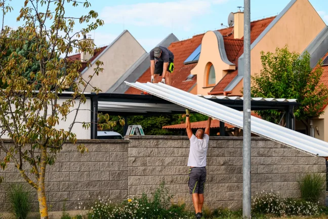 Pracovník podáva biely strešný panel kolegovi na streche prístrešku.