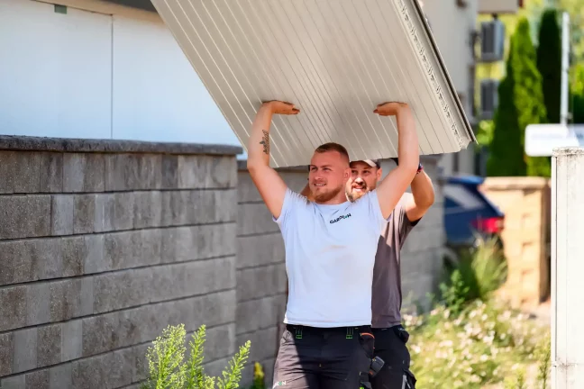 Dvaja pracovníci nesú nad hlavami veľký krémový strešný panel.
