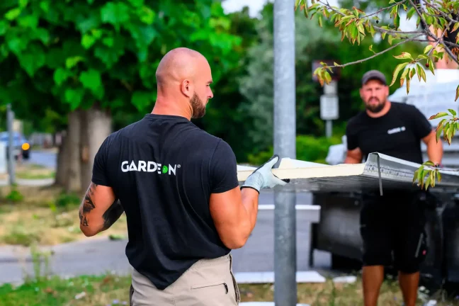 Dvaja pracovníci Gardeon nesú spoločne stenový panel popri ulici.
